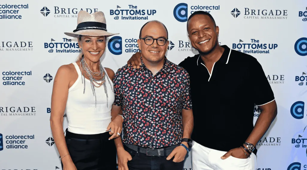 Three people smile for a photo at the Bottoms Up® Invitational step-and-repeat backdrop. The person on the left wears a white tank top, layered necklaces, black pants, and a wide-brimmed hat. The person in the middle wears glasses and a floral button-down shirt. The person on the right wears a black short-sleeve shirt with white trim and white pants. All appear happy and relaxed, standing close together in front of sponsor and event logos.