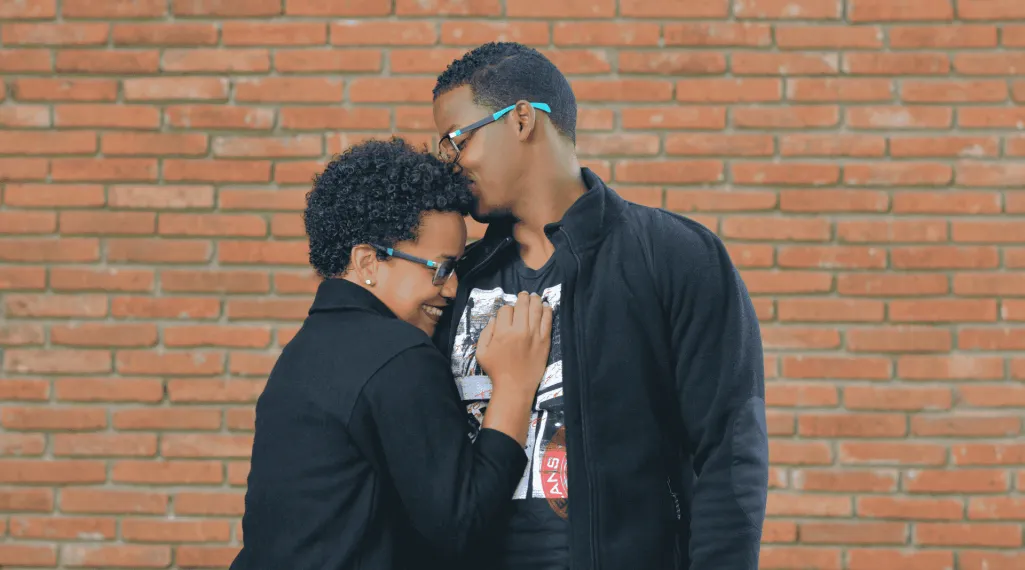 A smiling couple shares a quiet, affectionate moment in front of a brick wall, capturing warmth, connection, and everyday love.