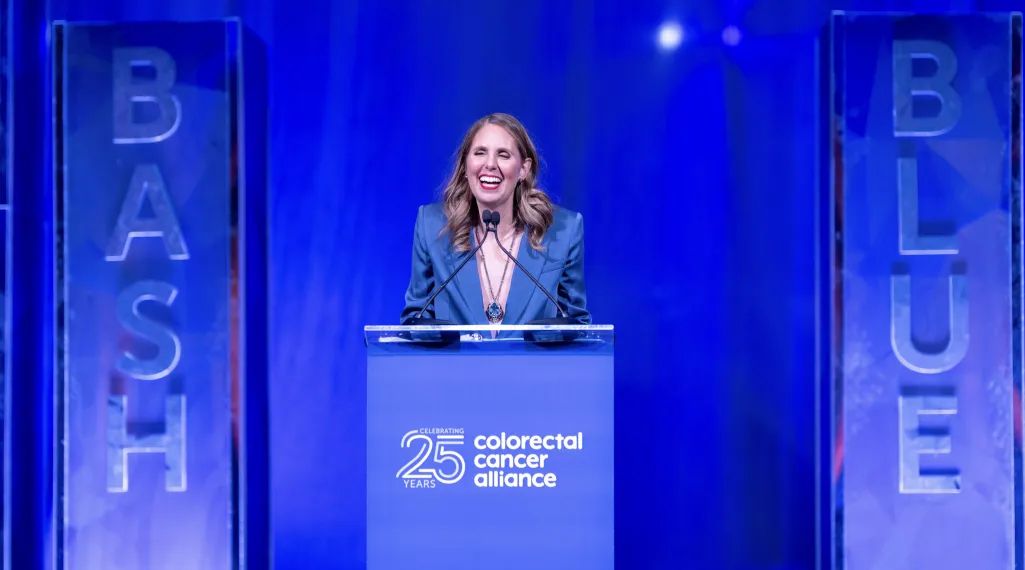 As event co-chair, Alison Friedman speaks at the podium during the Colorectal Cancer Alliance’s Blue Hope Bash in Washington, DC, in 2024. She smiles under bright stage lighting beside tall blue columns reading ‘BASH’ and ‘BLUE,’ with the Alliance's 25th anniversary logo displayed on the blue lectern.