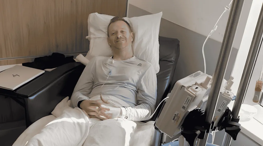 Andy smiles from a hospital bed, reclined against pillows with an IV pole and infusion pump beside him and a laptop on the tray table.