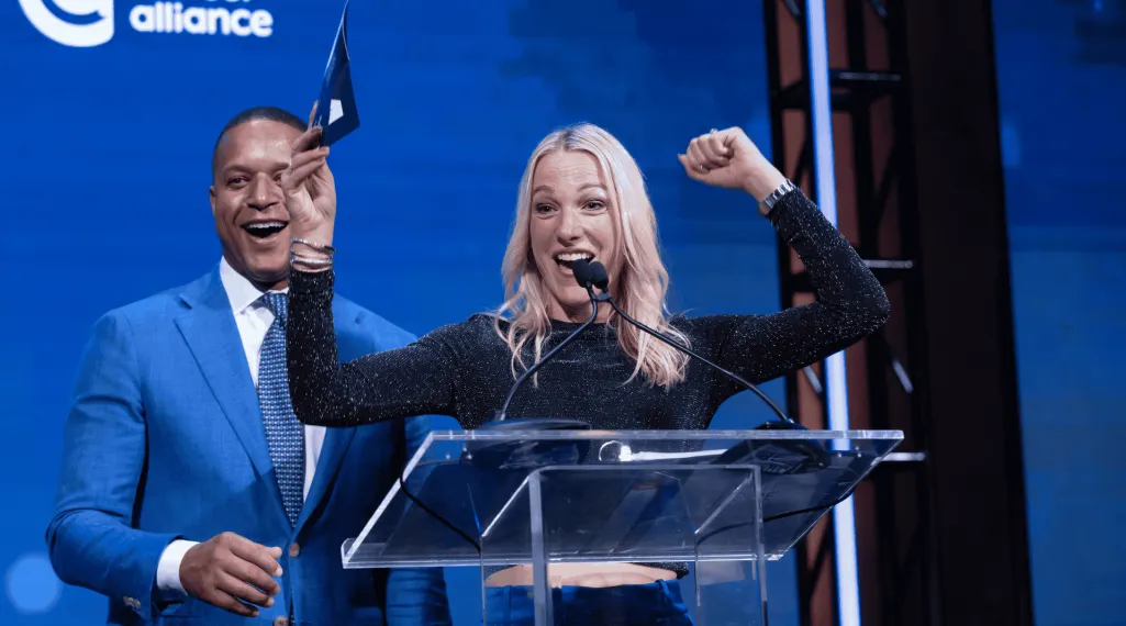 Craig Melvin and Lindsay Czarniak on stage at the Blue Hope Bash in Washington, DC. Lindsay stands at the podium smiling with one arm raised in celebration, while Craig, dressed in a blue suit, smiles beside her against a blue Colorectal Cancer Alliance backdrop.