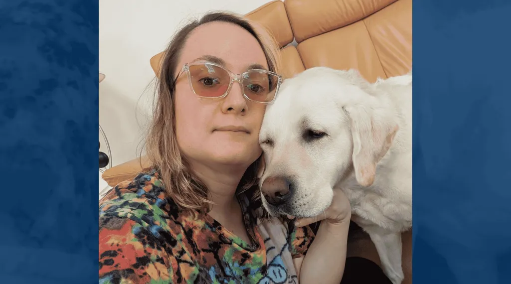 Jenny, wearing clear-framed glasses and a colorful tie-dye shirt, takes a selfie cheek to cheek with a white Labrador retriever on a tan leather couch.