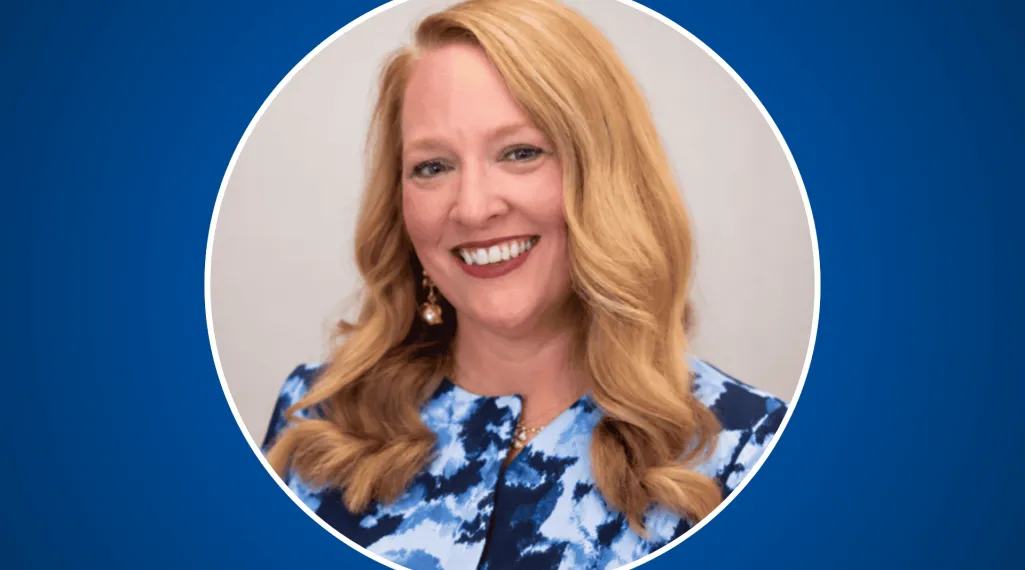 Headshot of Kate Watt, smiling, with long wavy strawberry-blonde hair, wearing a blue and white abstract-print top and pearl drop earrings, framed in a white-outlined circle on a royal blue background.