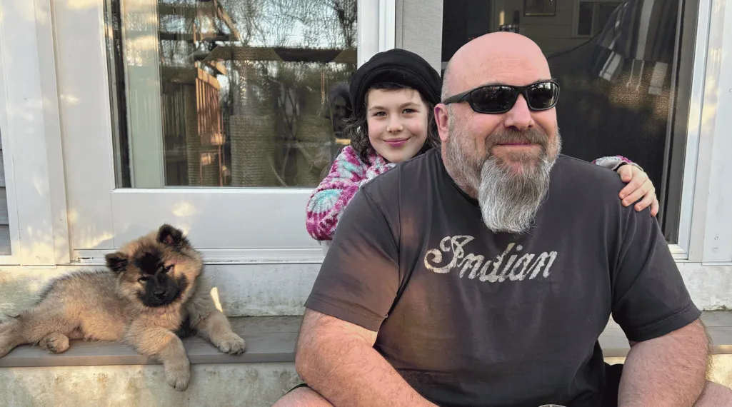 Kevin, a colorectal cancer patient, sits on a porch steps with one of his children, who rests their hands on his shoulders, and a fluffy puppy nearby.