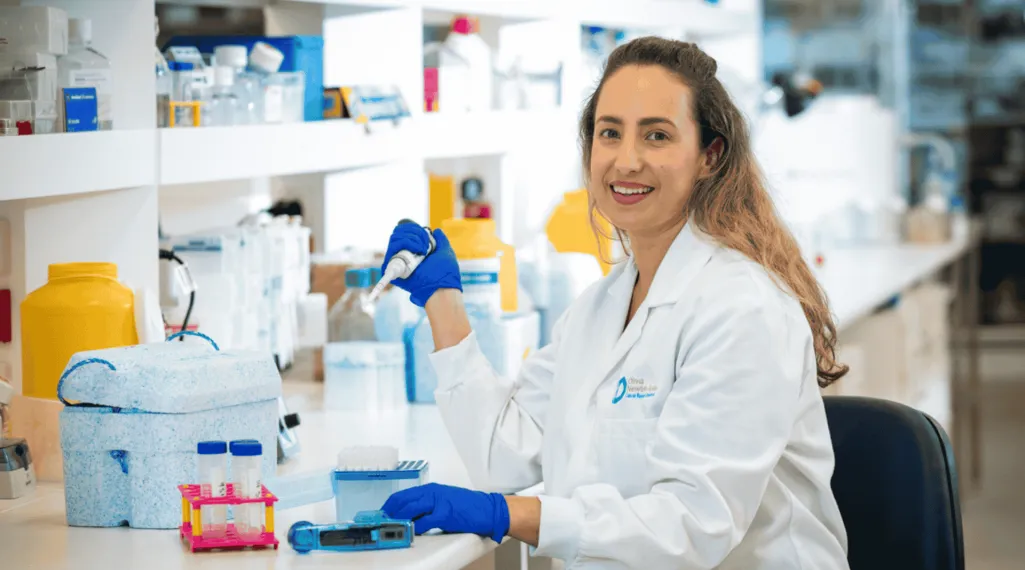 Dr. Mielke smiles while working in a research laboratory, wearing a white lab coat and blue gloves as she holds a pipette beside lab equipment.