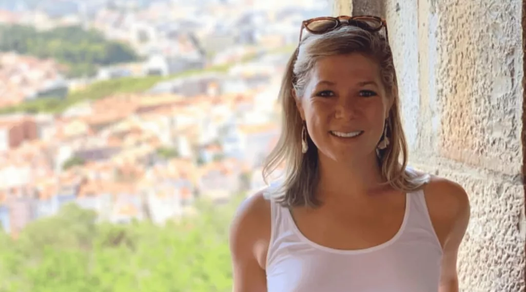A smiling woman with shoulder-length blonde hair stands in a stone archway, wearing a white tank top and sunglasses resting on her head. A blurred cityscape and greenery are visible in the background, suggesting a scenic viewpoint.