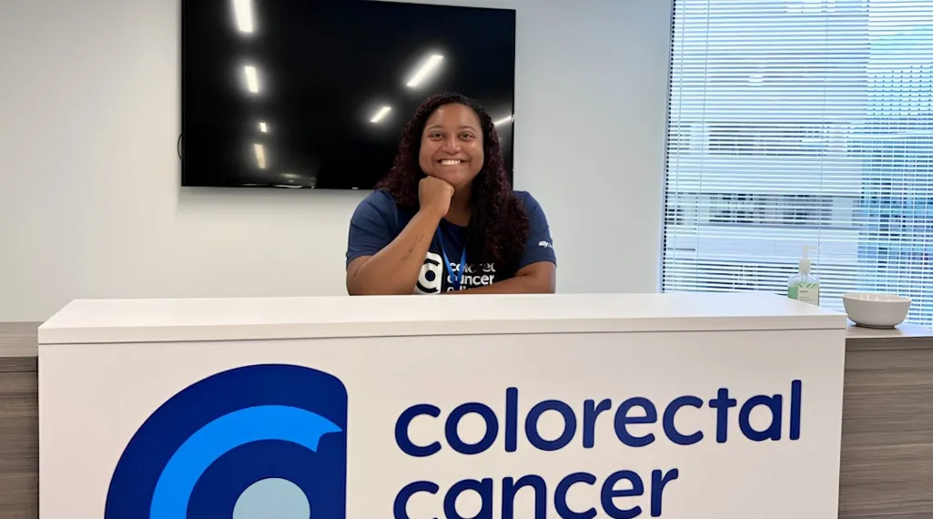 Paige Edmonds smiles while resting her chin on her hand at the Colorectal Cancer Alliance front desk, wearing an Alliance t-shirt and lanyard.