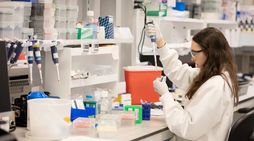A scientist at GSK's research center works in a lab coat, safety glasses, and gloves, using a pipette to transfer amber liquid into a small tube.