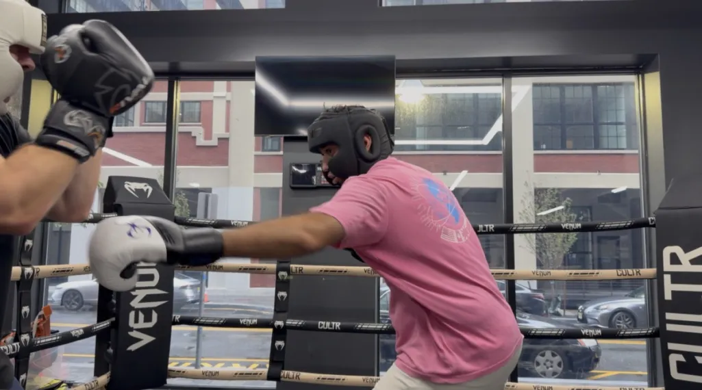 A boxer in a pink T-shirt and black headgear throws a punch with a white glove at a sparring partner in a boxing ring, with large windows behind them looking out on a city street.
