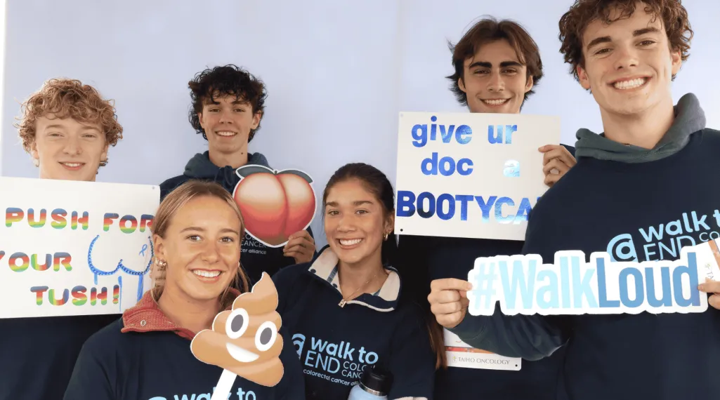 A group of smiling young people wearing “Walk to End Colon Cancer” shirts pose together holding playful awareness signs, including a peach emoji, a poop emoji, “Push for your tush!,” “Give ur doc a Bootycall,” and a “#WalkLoud” cutout.