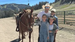 A smiling family of four, including John E., poses outdoors with a saddled brown horse at a mountain ranch, with pine-covered hills, fencing, and ski lift cables visible in the background on a sunny day.
