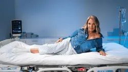 Katie Couric reclines confidently on a hospital bed wearing a denim jacket over patterned hospital pants, in a brightly lit medical setting with monitors and IV equipment visible in the background, promoting colon cancer screening awareness.
