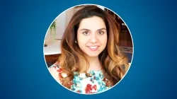 A smiling woman with long, wavy brown-to-blonde ombré hair looks at the camera. She is wearing a colorful floral top and standing indoors, with a softly blurred background of a room with wood floors and light decor. The portrait is cropped in a circular frame against a solid blue background.