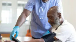 A healthcare worker wearing scrubs and gloves takes the blood pressure of an older man seated at a table.