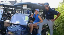 Craig Melvin and Lindsay Czarniak pose at a golf cart during The Bottoms Up Invitational, both wearing blue awareness ribbon apparel; the woman sits in the cart wearing a blue polo and visor, while the man stands beside her in a Bottoms Up Invitational cap and polo.
