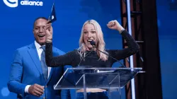 Craig Melvin and Lindsay Czarniak on stage at the Blue Hope Bash in Washington, DC. Lindsay stands at the podium smiling with one arm raised in celebration, while Craig, dressed in a blue suit, smiles beside her against a blue Colorectal Cancer Alliance backdrop.
