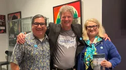 hree smiling people stand together indoors with their arms around each other. The person on the left wears glasses and a patterned shirt, the person in the middle wears a gray T-shirt and black blazer, and the person on the right wears glasses, a blue jacket, and a colorful scarf while holding a book. Behind them are framed artworks on the wall.
