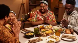 A family of four celebrates Kwanzaa, sitting around a festive dinner table with a lit kinara in the background, smiling and clapping hands together.