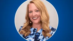 Headshot of Kate Watt, smiling, with long wavy strawberry-blonde hair, wearing a blue and white abstract-print top and pearl drop earrings, framed in a white-outlined circle on a royal blue background.