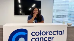 Paige Edmonds smiles while resting her chin on her hand at the Colorectal Cancer Alliance front desk, wearing an Alliance t-shirt and lanyard.