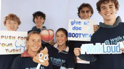A group of smiling young people wearing “Walk to End Colon Cancer” shirts pose together holding playful awareness signs, including a peach emoji, a poop emoji, “Push for your tush!,” “Give ur doc a Bootycall,” and a “#WalkLoud” cutout.