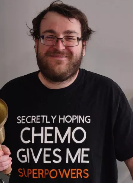 Man wearing "Secretly hoping chemo gives me superpowers" shirt holding a big bell