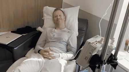 Andy smiles from a hospital bed, reclined against pillows with an IV pole and infusion pump beside him and a laptop on the tray table.