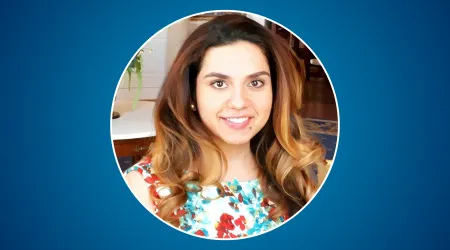 A smiling woman with long, wavy brown-to-blonde ombré hair looks at the camera. She is wearing a colorful floral top and standing indoors, with a softly blurred background of a room with wood floors and light decor. The portrait is cropped in a circular frame against a solid blue background.