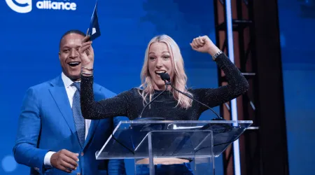 Craig Melvin and Lindsay Czarniak on stage at the Blue Hope Bash in Washington, DC. Lindsay stands at the podium smiling with one arm raised in celebration, while Craig, dressed in a blue suit, smiles beside her against a blue Colorectal Cancer Alliance backdrop.