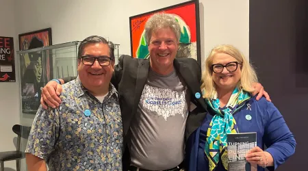 hree smiling people stand together indoors with their arms around each other. The person on the left wears glasses and a patterned shirt, the person in the middle wears a gray T-shirt and black blazer, and the person on the right wears glasses, a blue jacket, and a colorful scarf while holding a book. Behind them are framed artworks on the wall.