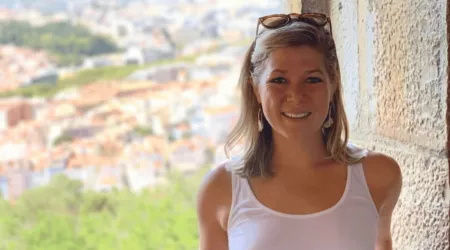 A smiling woman with shoulder-length blonde hair stands in a stone archway, wearing a white tank top and sunglasses resting on her head. A blurred cityscape and greenery are visible in the background, suggesting a scenic viewpoint.