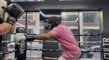 A boxer in a pink T-shirt and black headgear throws a punch with a white glove at a sparring partner in a boxing ring, with large windows behind them looking out on a city street.