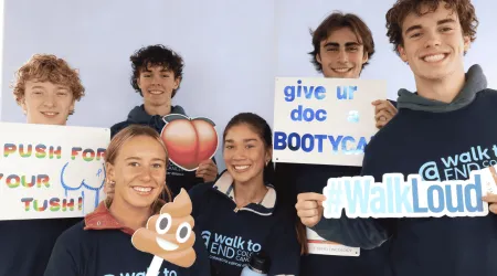 A group of smiling young people wearing “Walk to End Colon Cancer” shirts pose together holding playful awareness signs, including a peach emoji, a poop emoji, “Push for your tush!,” “Give ur doc a Bootycall,” and a “#WalkLoud” cutout.