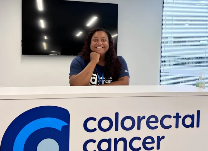 Paige Edmonds smiles while resting her chin on her hand at the Colorectal Cancer Alliance front desk, wearing an Alliance t-shirt and lanyard.