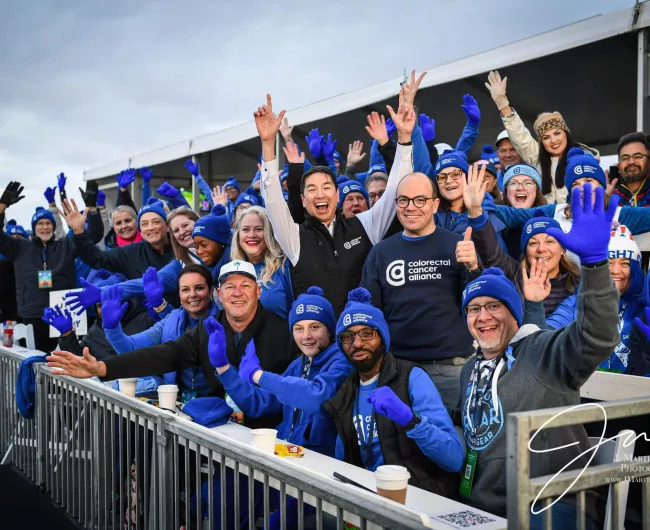 An energetic group of people at an event, the Cologuard Classic golf tournament, many wearing Colorectal Cancer Alliance gear and blue gloves, cheering and smiling toward the camera. 