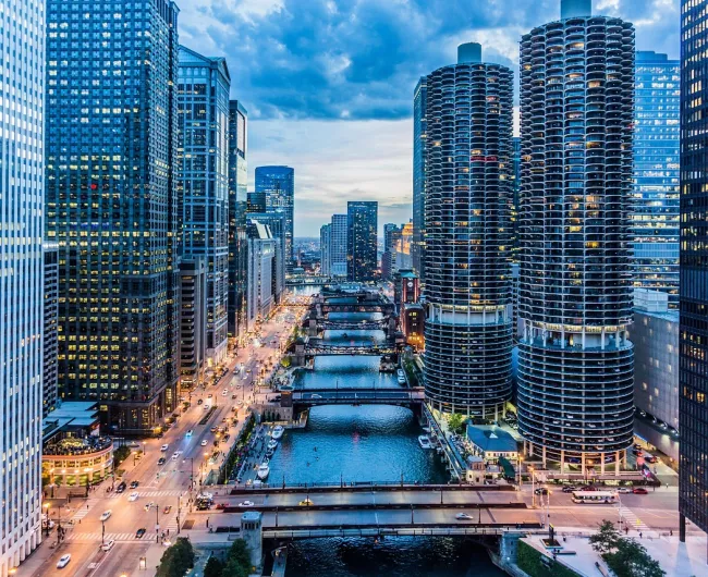 A vibrant aerial view of downtown Chicago at dusk, featuring the illuminated skyscrapers along the Chicago River, several bridges crossing the water, and a lively street scene below under a partly cloudy sky.