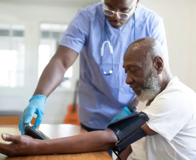 A healthcare worker wearing scrubs and gloves takes the blood pressure of an older man seated at a table.