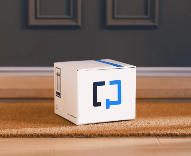 A Cologuard test kit box sitting on a woven doormat at a residential front door, with "this side up" arrows and a "Human Specimen" label printed on the box.