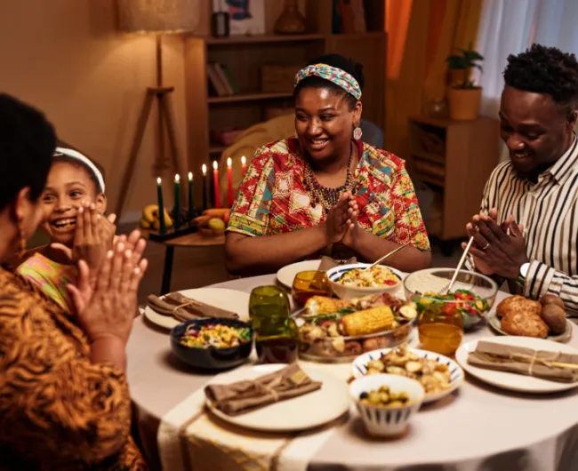 A family of four celebrates Kwanzaa, sitting around a festive dinner table with a lit kinara in the background, smiling and clapping hands together.
