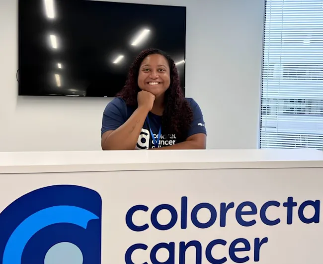 Paige Edmonds smiles while resting her chin on her hand at the Colorectal Cancer Alliance front desk, wearing an Alliance t-shirt and lanyard.