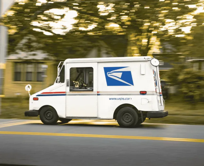 A USPS mail truck driving down a tree-lined residential street, shown with motion blur suggesting it's moving quickly along its route.