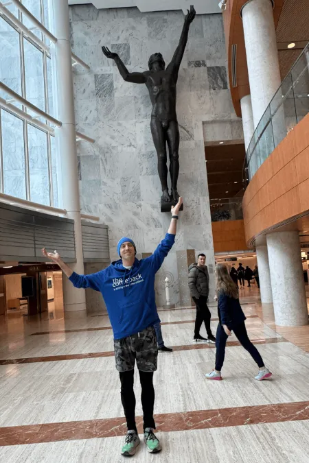 John E., wearing a blue hoodie, stands inside a large, light-filled atrium, smiling and raising his arms to mirror a tall bronze sculpture of a human figure with uplifted arms suspended above him, while people walk through the space behind him.