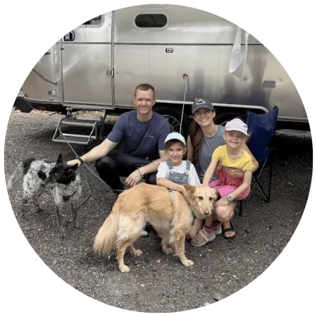 Andy crouches beside his wife and two daughters in front of a silver Airstream trailer, with two dogs in the foreground.