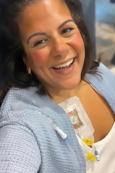 A smiling woman wearing a light blue cardigan and white top takes a selfie. A medical port and tubing are visible on her upper chest, secured with a transparent dressing. She appears cheerful and is looking directly at the camera.