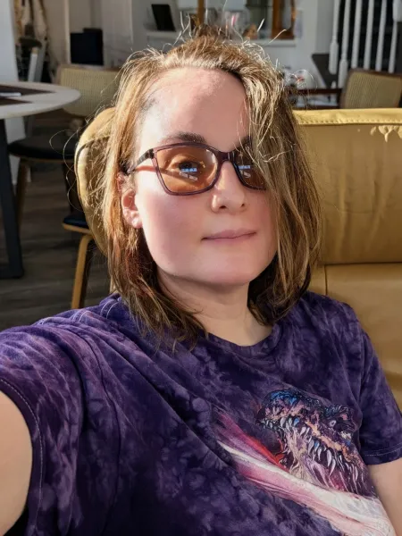 A sunlit selfie of Jenny in a yellow leather chair, wearing amber-tinted glasses and a purple dragon T-shirt, with a soft smile.