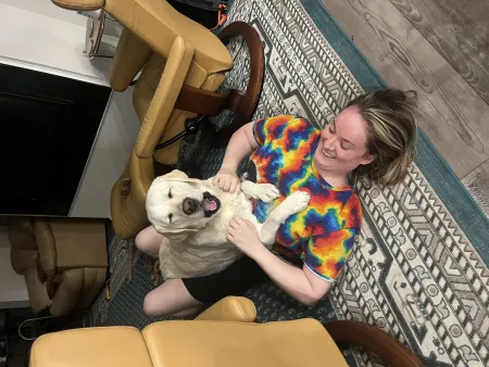 Jenny laughs on the living room floor while a happy cream-colored Labrador sits on her chest, grinning.