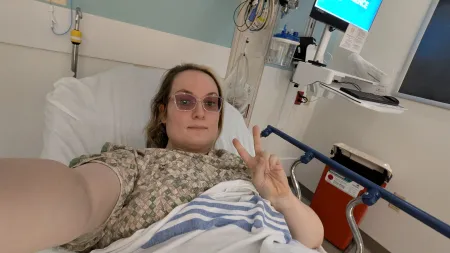 Jenny in a hospital bed and gown, flashing a peace sign at the camera with a calm, slight smile.