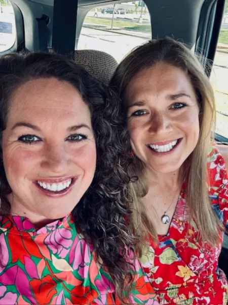 Two smiling women sit side by side in a car, wearing bright, floral-patterned dresses. One has curly dark hair, and the other has straight blonde hair. Sunlight filters in through the windows behind them, creating a cheerful, warm atmosphere.