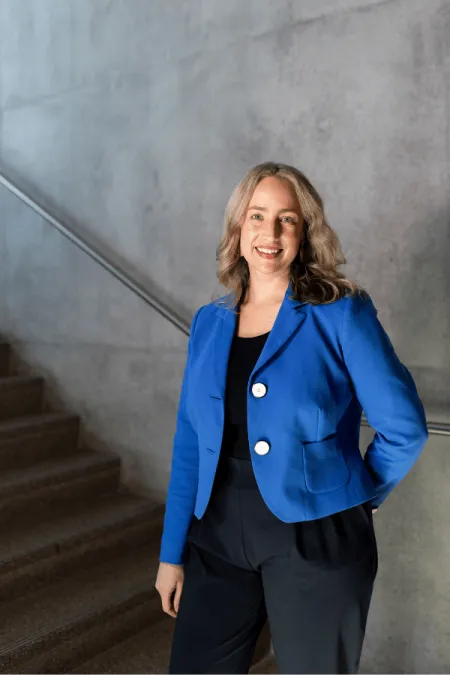 Dr. Mielke stands indoors wearing a blue blazer and black outfit, smiling confidently against a modern concrete backdrop.