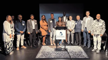 Twelve speakers and panelists pose together on a dimly lit stage at the ScreenSmart event, smiling behind a podium with a sign reading "ScreenSmart: data | access | adherence." Folding chairs line the stage behind them, and a blue patterned rug sits in front of the podium.